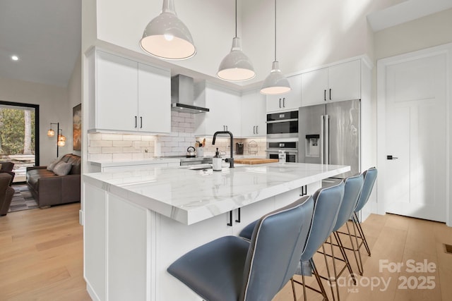 kitchen with decorative light fixtures, wall chimney range hood, decorative backsplash, stainless steel appliances, and white cabinets