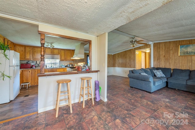 kitchen with ceiling fan, a breakfast bar area, a textured ceiling, kitchen peninsula, and wall chimney exhaust hood