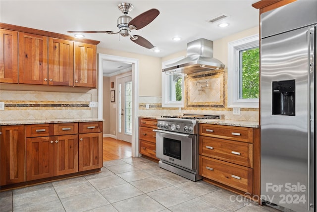 kitchen with light stone counters, ceiling fan, wall chimney exhaust hood, high quality appliances, and backsplash