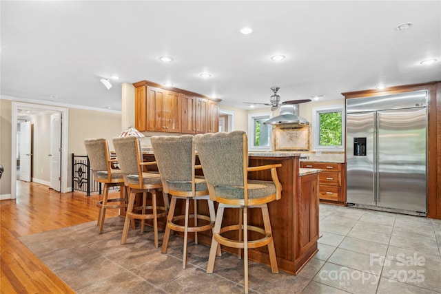 kitchen featuring built in refrigerator, a ceiling fan, brown cabinets, light stone countertops, and tasteful backsplash