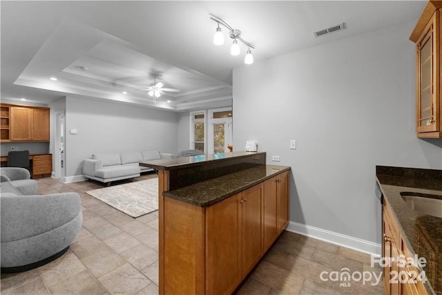 kitchen featuring kitchen peninsula, ceiling fan, light tile floors, dark stone counters, and a tray ceiling