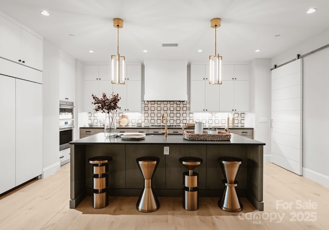 kitchen with premium range hood, pendant lighting, an island with sink, a barn door, and white cabinetry