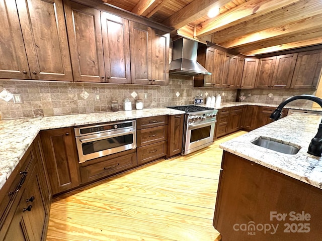 kitchen featuring beamed ceiling, appliances with stainless steel finishes, light stone countertops, and wall chimney range hood