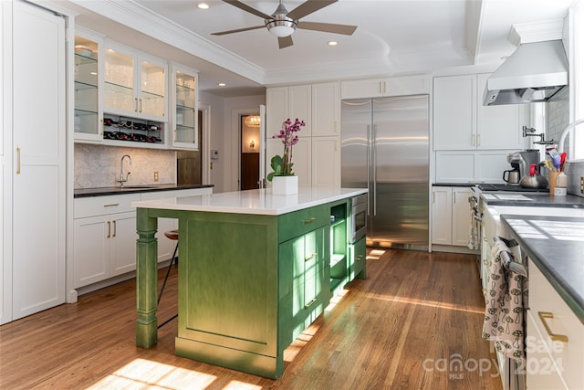 kitchen with a kitchen island, wall chimney range hood, built in appliances, light hardwood / wood-style flooring, and white cabinetry