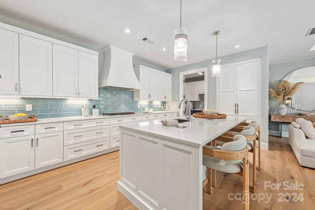 kitchen with a center island with sink, pendant lighting, light hardwood / wood-style floors, white cabinetry, and custom exhaust hood