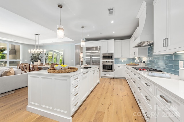 kitchen featuring white cabinetry, decorative light fixtures, premium range hood, a center island with sink, and light hardwood / wood-style floors