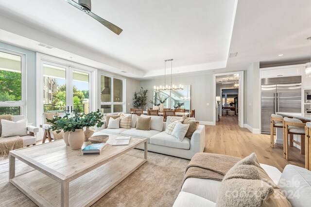 living room with ceiling fan with notable chandelier, a raised ceiling, and light hardwood / wood-style flooring