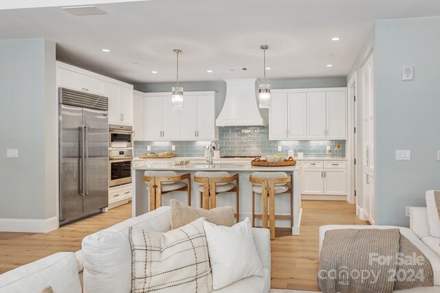 kitchen with pendant lighting, light hardwood / wood-style flooring, stainless steel appliances, premium range hood, and an island with sink