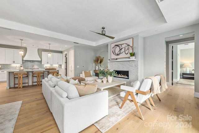 living room featuring a raised ceiling, a fireplace, sink, ceiling fan, and light wood-type flooring