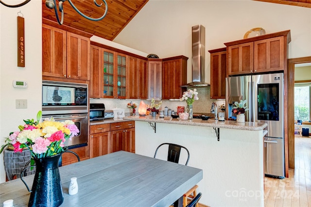 kitchen with wall chimney range hood, stainless steel appliances, decorative backsplash, light stone counters, and a breakfast bar