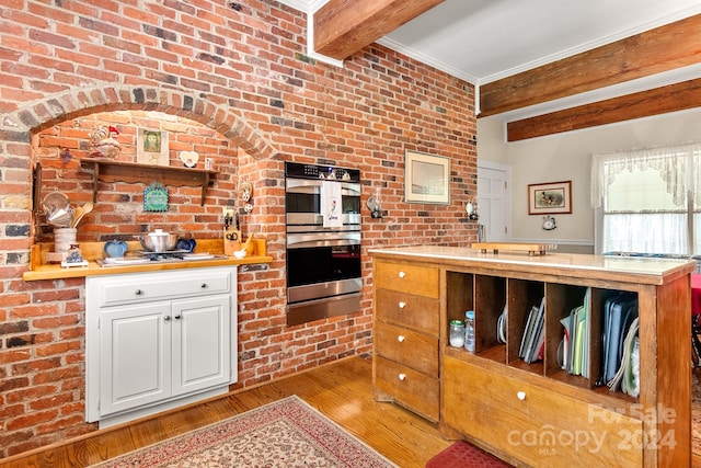 kitchen featuring beamed ceiling, stainless steel double oven, white cabinets, and brick wall