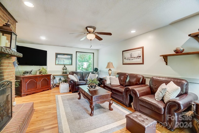 living room with ceiling fan, light wood-type flooring, a brick fireplace, and a textured ceiling