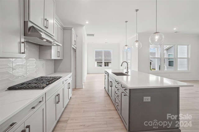 kitchen with stainless steel appliances, a kitchen island with sink, sink, pendant lighting, and light hardwood / wood-style flooring