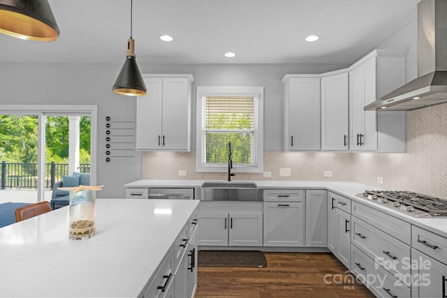 kitchen with white cabinets, wall chimney range hood, stainless steel appliances, sink, and hanging light fixtures