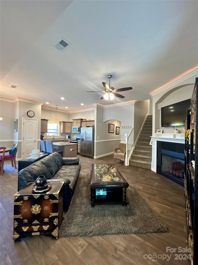 living room with ceiling fan, a tiled fireplace, dark wood-type flooring, sink, and ornamental molding