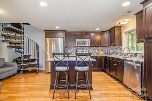 kitchen with appliances with stainless steel finishes, a kitchen island, backsplash, light wood-type flooring, and light stone countertops