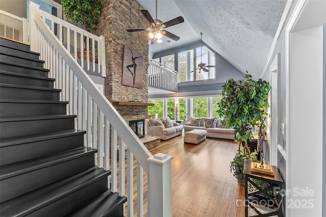 living room with ceiling fan, hardwood / wood-style flooring, a stone fireplace, a textured ceiling, and high vaulted ceiling