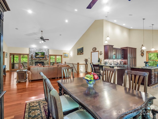 dining space with high vaulted ceiling, ceiling fan, a stone fireplace, and light hardwood / wood-style flooring