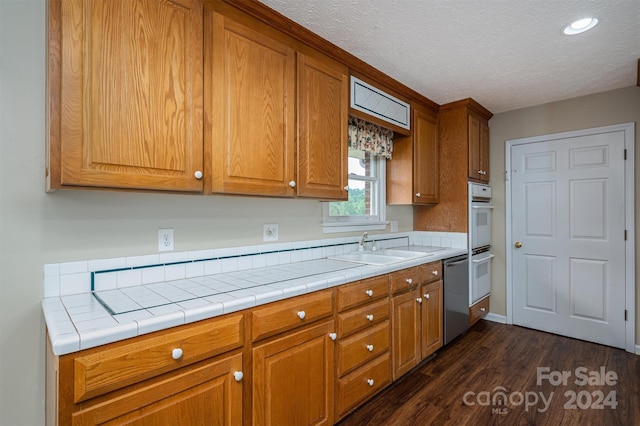 kitchen featuring dark wood-type flooring, stainless steel dishwasher, sink, tile countertops, and a textured ceiling