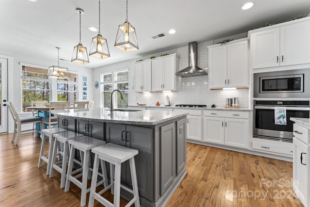 kitchen with appliances with stainless steel finishes, wall chimney range hood, an island with sink, and light hardwood / wood-style flooring