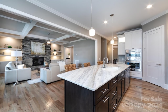 kitchen featuring stainless steel double oven, a kitchen island with sink, sink, white cabinets, and beamed ceiling