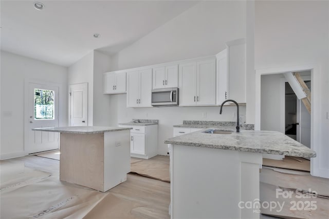 kitchen with white cabinets, a sink, stainless steel microwave, light stone counters, and high vaulted ceiling
