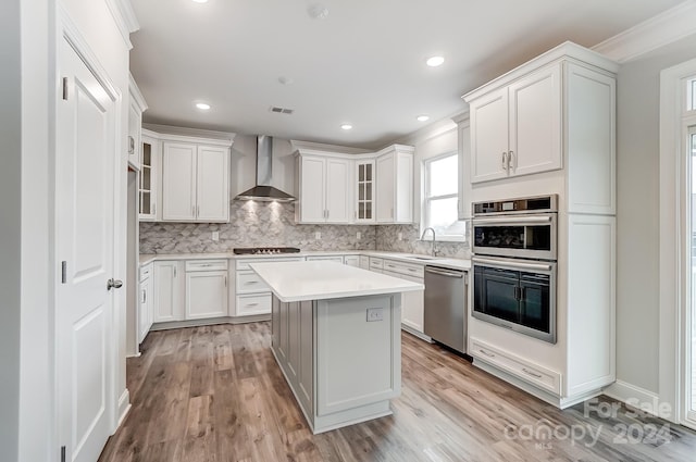 kitchen with a center island, wall chimney exhaust hood, appliances with stainless steel finishes, light hardwood / wood-style floors, and white cabinetry