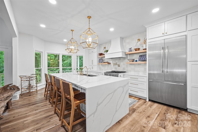 kitchen with built in refrigerator, light stone counters, custom range hood, white cabinets, and a kitchen bar