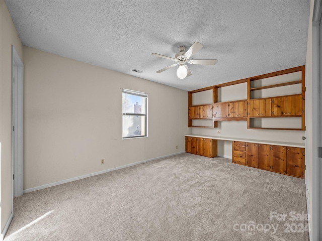 unfurnished living room with light colored carpet, a textured ceiling, built in desk, and ceiling fan