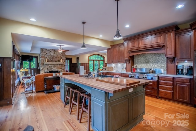 kitchen with a stone fireplace, backsplash, hanging light fixtures, a kitchen island with sink, and butcher block countertops