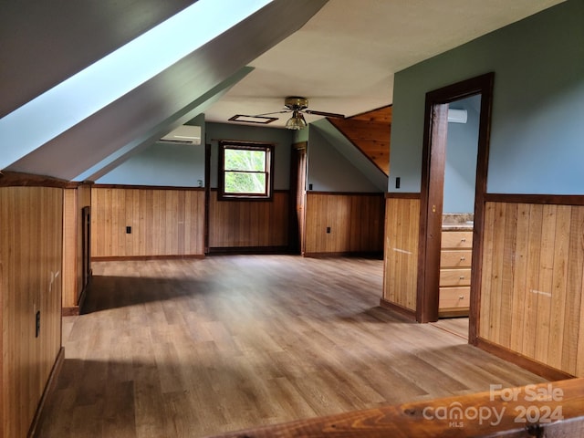 bonus room with an AC wall unit, vaulted ceiling with skylight, wooden walls, light wood-type flooring, and ceiling fan