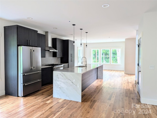 kitchen with high end fridge, a center island with sink, pendant lighting, wall chimney range hood, and backsplash