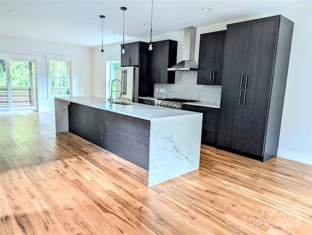 kitchen with wall chimney range hood, light hardwood / wood-style flooring, backsplash, a center island with sink, and decorative light fixtures