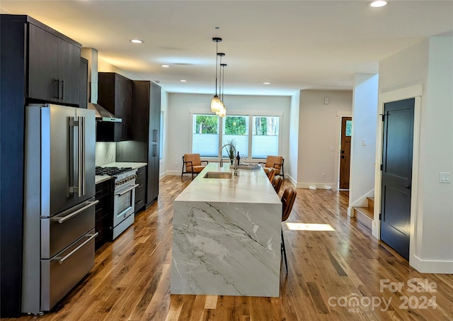 kitchen with sink, high end appliances, an island with sink, light stone countertops, and wall chimney range hood