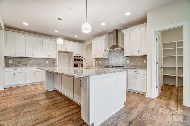 kitchen with decorative light fixtures, backsplash, an island with sink, white cabinets, and wall chimney exhaust hood