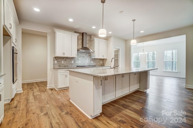 kitchen featuring light stone countertops, pendant lighting, wall chimney exhaust hood, white cabinetry, and an island with sink