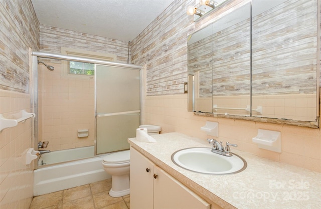 full bathroom with vanity, bath / shower combo with glass door, toilet, a textured ceiling, and tile walls