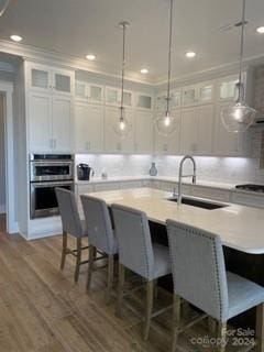 kitchen with stainless steel double oven, decorative light fixtures, white cabinetry, sink, and a kitchen island with sink