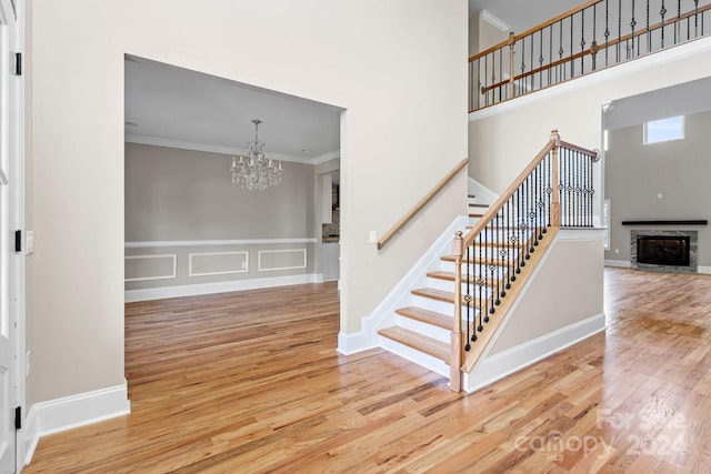 staircase featuring a high ceiling, a stone fireplace, a notable chandelier, hardwood / wood-style flooring, and ornamental molding