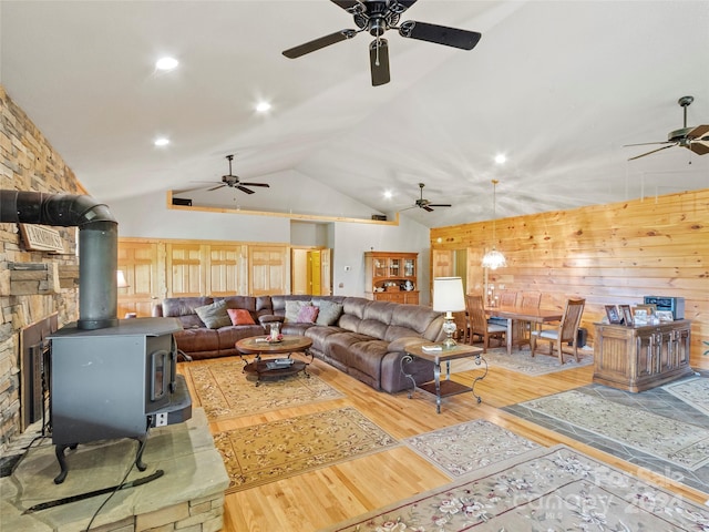 living room featuring wooden walls, ceiling fan, a wood stove, light wood-type flooring, and vaulted ceiling