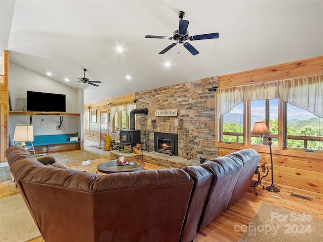 living room with wood walls, ceiling fan, a wood stove, lofted ceiling, and light hardwood / wood-style flooring