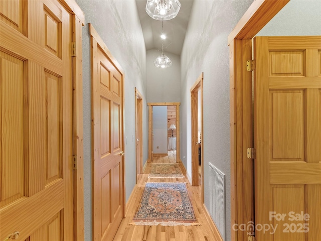 corridor featuring visible vents, a textured wall, a towering ceiling, a chandelier, and light wood-type flooring