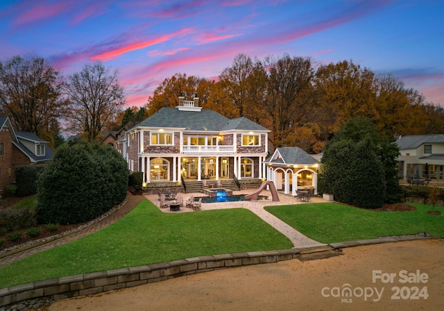 back house at dusk with a balcony, a patio area, and a lawn