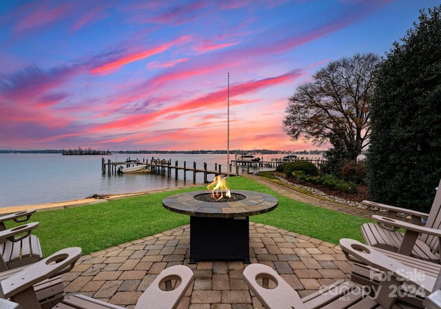 patio terrace at dusk featuring a boat dock, a yard, a water view, and an outdoor fire pit