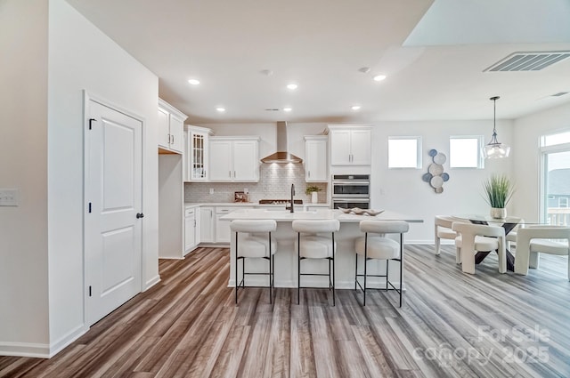 kitchen featuring pendant lighting, wall chimney range hood, white cabinetry, a center island with sink, and light wood-type flooring