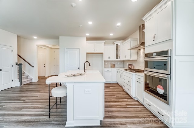 kitchen featuring white cabinetry, wall chimney exhaust hood, stainless steel appliances, and a center island with sink