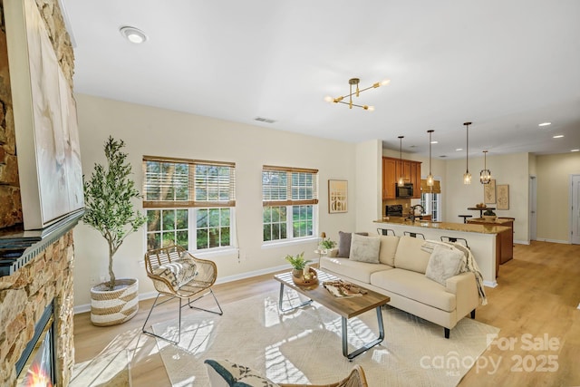 living room featuring a stone fireplace, visible vents, baseboards, light wood-style floors, and an inviting chandelier