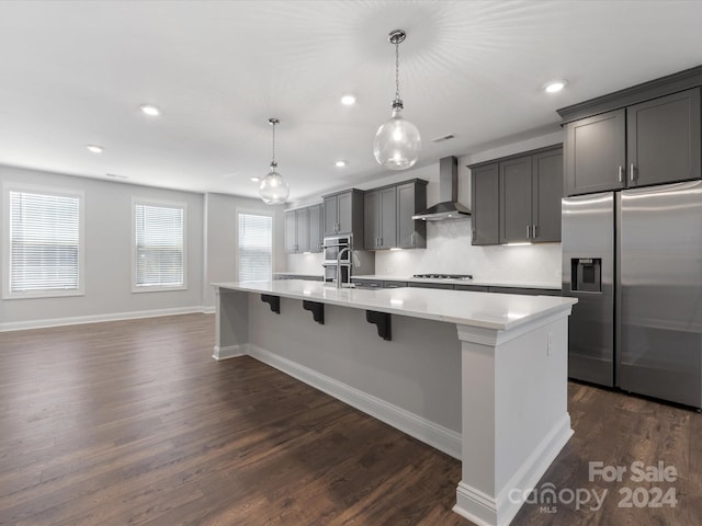 kitchen with wall chimney exhaust hood, stainless steel appliances, dark wood-type flooring, decorative light fixtures, and a center island with sink