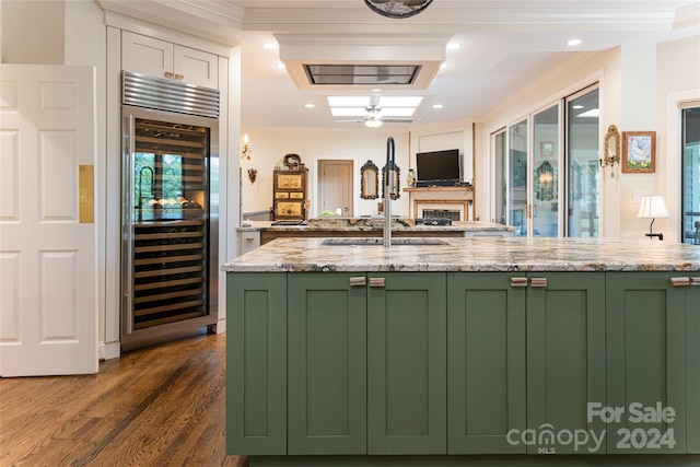 kitchen with green cabinets, crown molding, wine cooler, light stone countertops, and an island with sink