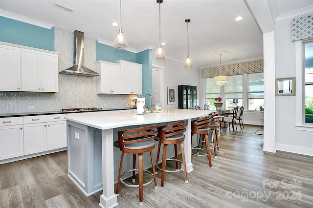 kitchen featuring decorative light fixtures, wall chimney exhaust hood, white cabinets, an island with sink, and ornamental molding